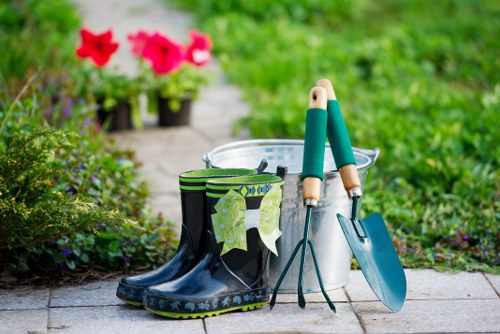 A garden scene featuring a paved pathway with a pair of black rubber gardening boots placed on the ground, one of which has a pair of green and white gardening gloves draped over it. To the right of the boots, there is a small metal trowel and a hand fork with green handles resting on a white plastic bucket, which is partially visible. In the background, there are vibrant pink and red flowers in pots, set amidst lush green foliage and grass, creating a colourful and well-maintained outdoor area typical of a residential garden in Norbiton. The scene suggests preparations or ongoing maintenance work, with a focus on planting and outdoor care, reflecting professional gardening and landscaping services. The sunlight indicates a bright, clear day with natural light illuminating the scene, emphasizing the natural tones of the garden elements and the well-kept environment.