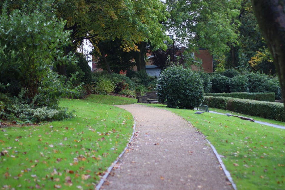A gardener wearing a dark green uniform with yellow stripe accents, labeled 'SAIGON ZOO' on the back, walks along a paved pathway in a lush, landscaped outdoor area. The gardener is carrying a blue sp